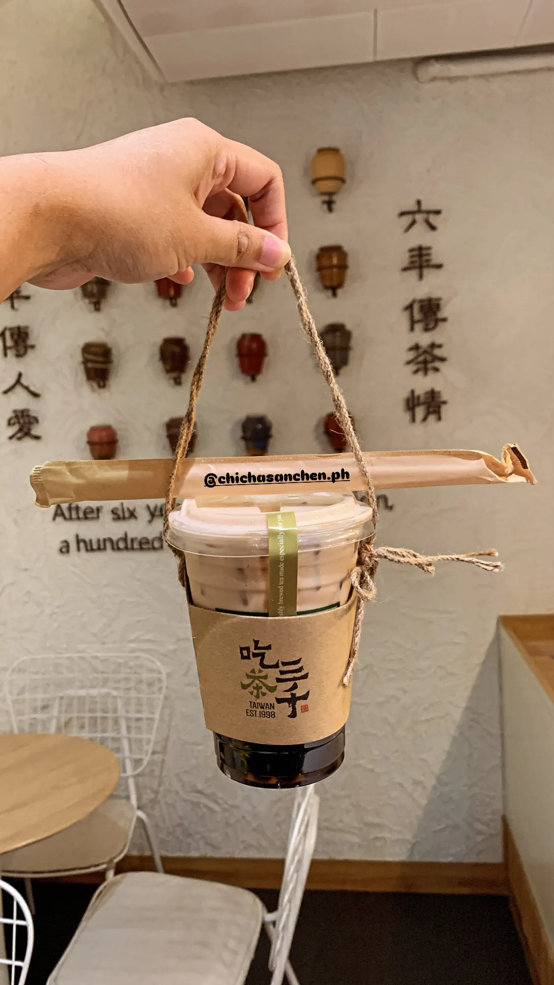 A cup of bubble milk tea hanging by a twine string held by my hand. A white brick wall with colorful ceramic teapots are in the background.