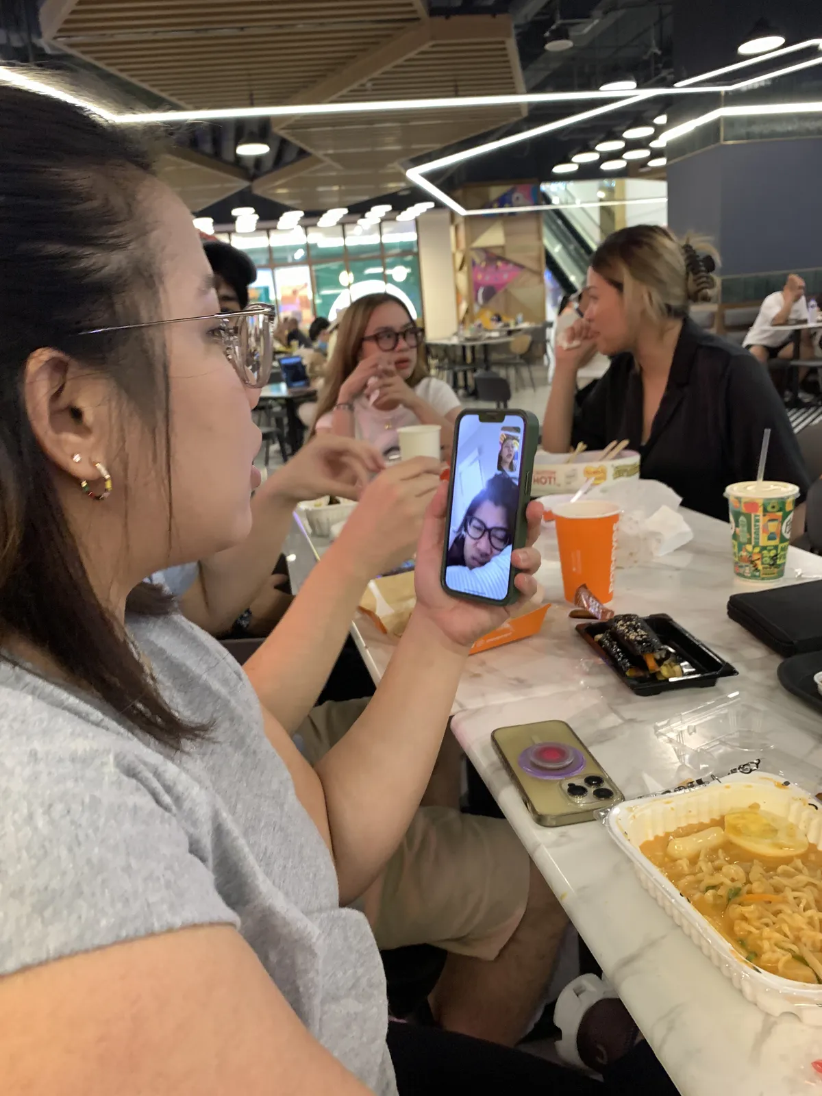 A couple of young women seated at one end of a long table at the food court. The woman on the foreground is holding a phone with another woman on videocall.