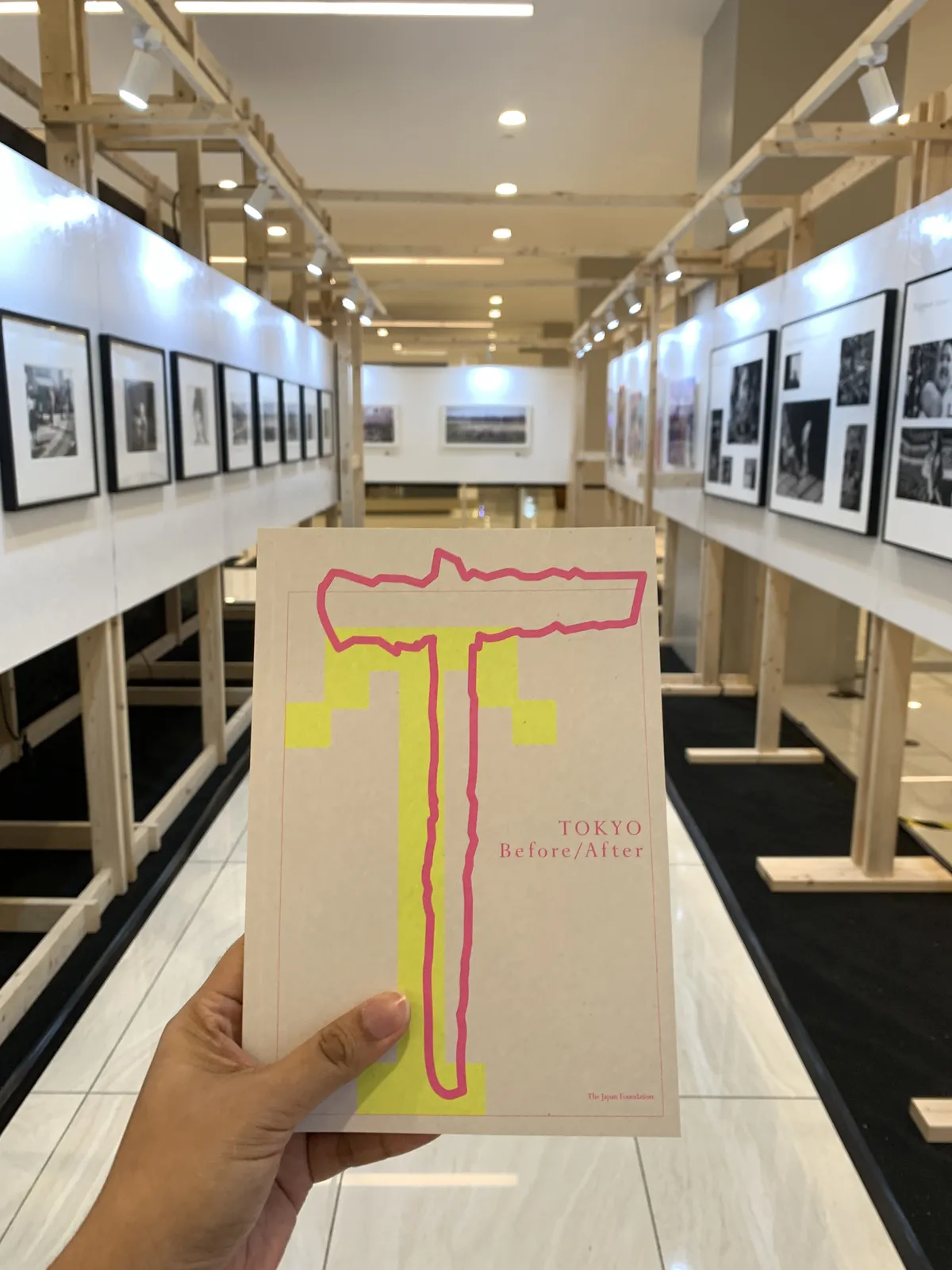 A hand holding a small guidebook for an exhibit titled Tokyo Before/After. In the background is a hallway of framed black-and-white photographs.