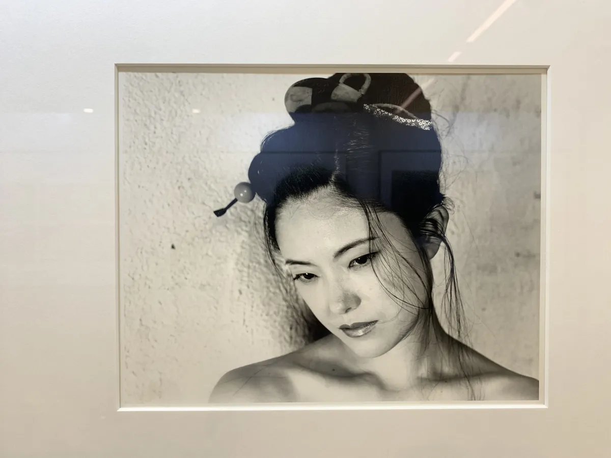 A black and white photograph of a Japanese woman from her shoulders up, with striking features and collarbones that look like sand dunes. She is topless and her hair is in a messy geisha bun, wisps of hair falling to her face.