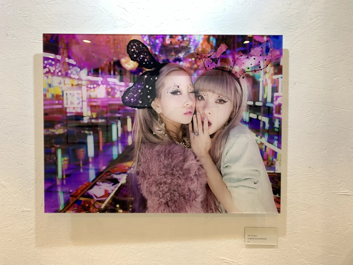 A photo of two women in rabbit ears and heavy makeup looking into the camera. One woman is winking and the other is feigning surprise, hand covering her mouth. They are in a room lit up with colorful neon lights.