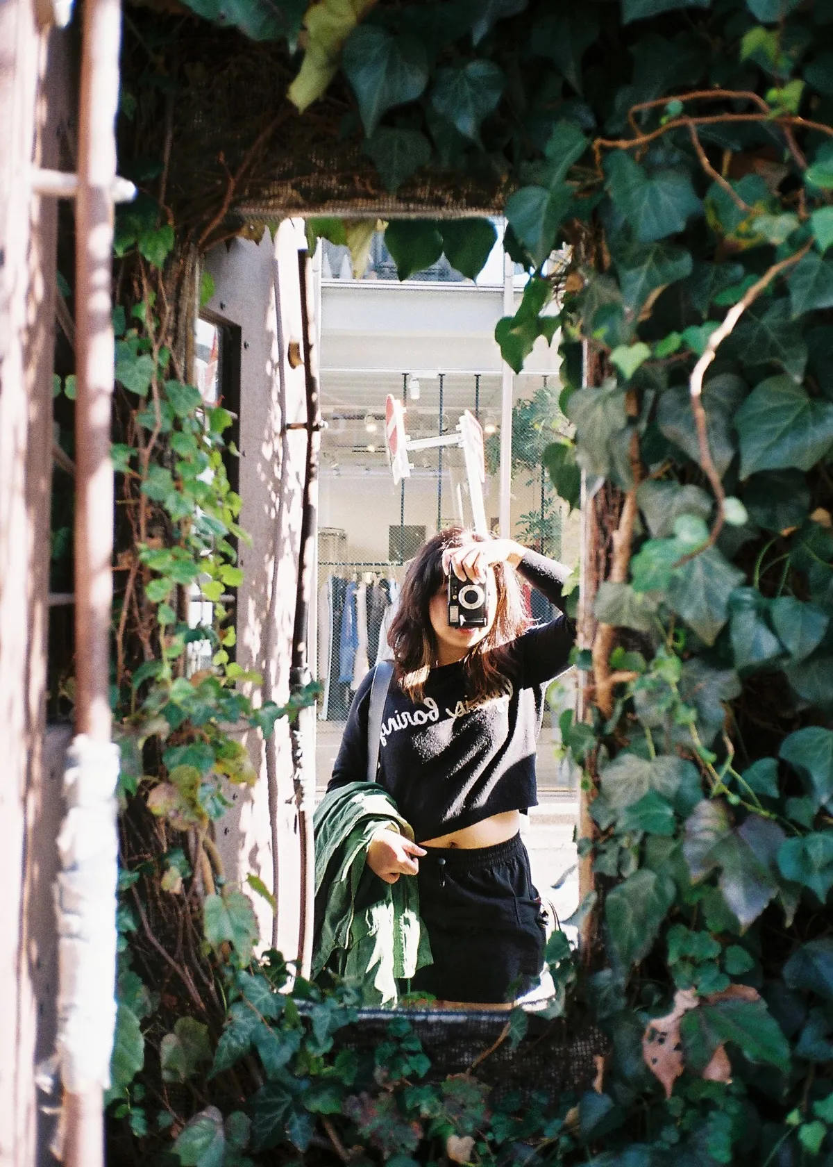 A woman wearing a black sweater riding up her tummy and a black skirt taking a selfie on a wall mirror nestled with plants outside. A Konica Genbakantoku film camera obscures her face.