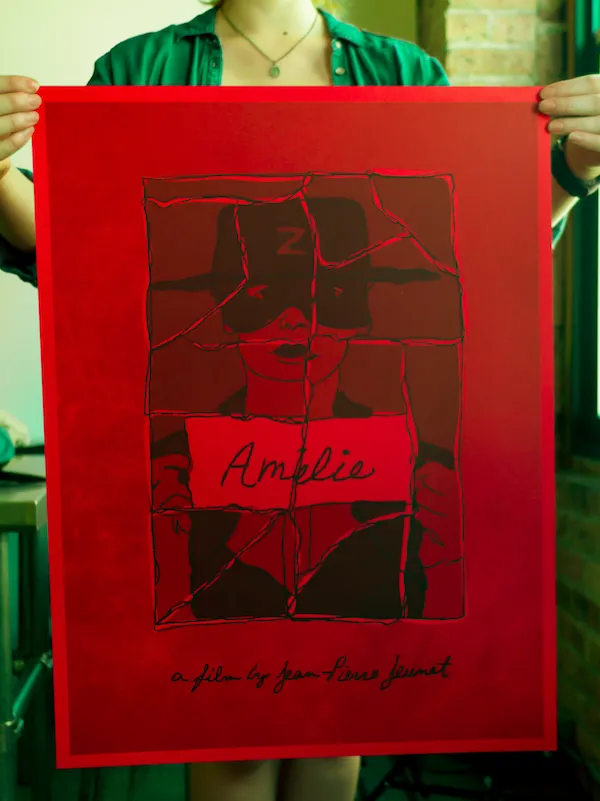 A person holding a red art poster featuring a hand-drawn illustration of Amélie in an eye mask and hat, overlaid with a shattered glass pattern and the title 'Amélie'.