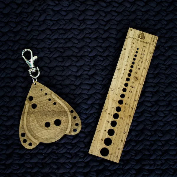 A photo of custom wooden knitting tools resting on a dark blue, chunky knit background: a teardrop-shaped needle gauge shaped like the firefly-yarn logo with a keychain clasp, and a rectangular wooden ruler featuring measurement markings and the