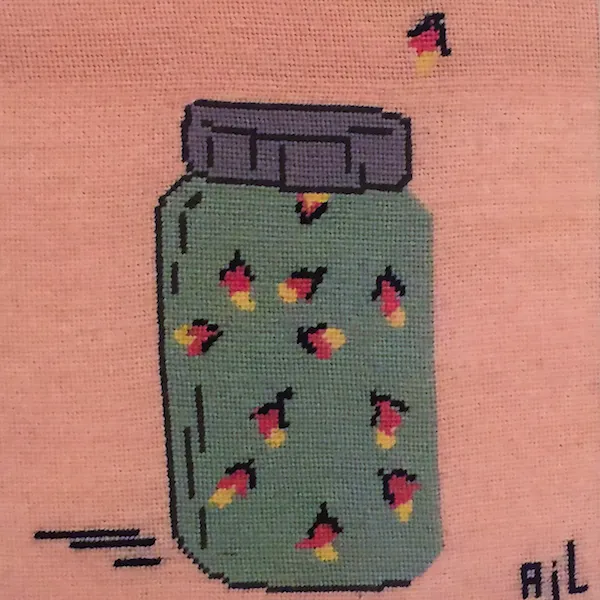 A cross-stitch piece on pink fabric showing a mason jar filled with fireflies. The jar has a grey lid and a teal body containing several stitched fireflies with pink bodies and yellow glowing tails, with one stray firefly floating outside above the jar.