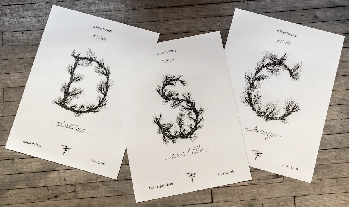 A flat lay photograph of three white concert tour posters for 'A Fine Frenzy PINES' arranged on a rustic wooden surface. Each poster features minimalist typography at the top, a large central letter formed out of detailed black ink illustrations of pine tree branches, the city name in a script font at the bottom, and a small date and venue footer. The letters shown are 'D' for Dallas on the left, 'S' for Seattle in the center, and 'C' for Chicago on the right.