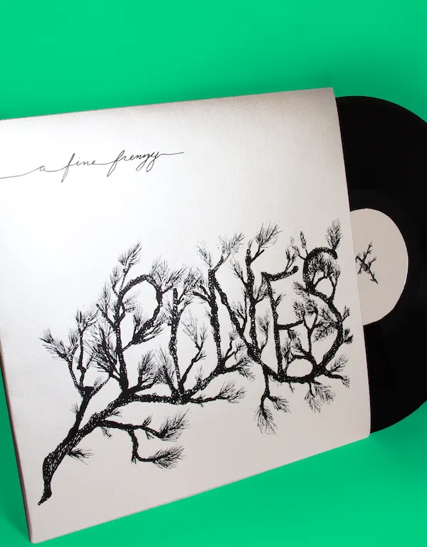 An album cover of 'Pines' vinyl package by A Fine Frenzy set against a vibrant green background. The off-white jacket features an intricate black ink illustration where the word 'Pines' is formed entirely out of textured pine tree branches, with 'A Fine Frenzy' written in a delicate script at the top left. A black vinyl record with a white center label is partially pulled out from the right side of the sleeve.