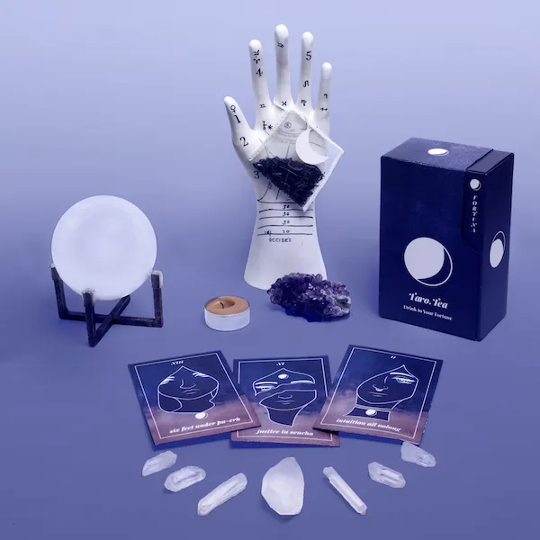 A product arrangement for 'Taro·Tea' on a purple surface. Three tarot-style tea packaging cards labeled 'six feet under pu-erh', 'justice in sencha', and 'intuition all oolong' are laid out in front of a dark purple box. An arc of small quartz crystals sits below the cards, while a ceramic palmistry hand, crystal ball, amethyst cluster, and small candle are arranged in the background.