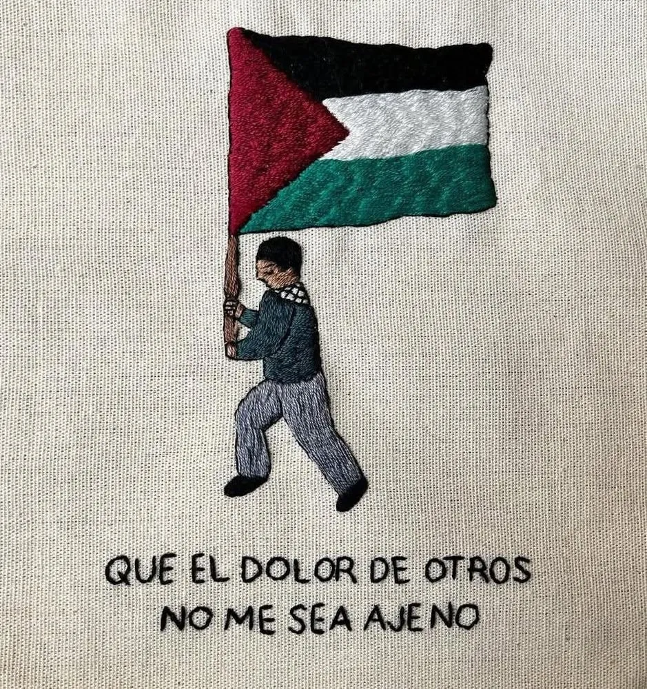 Fonte: internet. Foto de um tecido pontilhado em fundo claro, contendo uma pessoa de cabelo curto, pele marrom, brusa verde, calça cinza, sapato preto, segurando uma bandeira da Palestina, e embaixo da pessoa tem costurada a frase em espanhol “Que a dor de outros não me seja alheia”