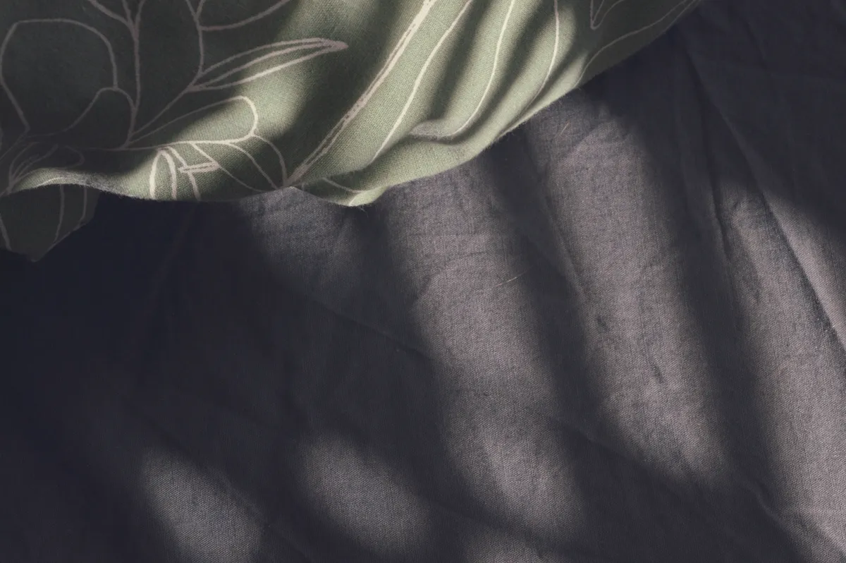 A horizontal colour photograph of a corner of a bed unmade. The sheet is a dark grey. The duvet cover is a white and green leafy pattern. Light streaks in horizontal lines over the scene.