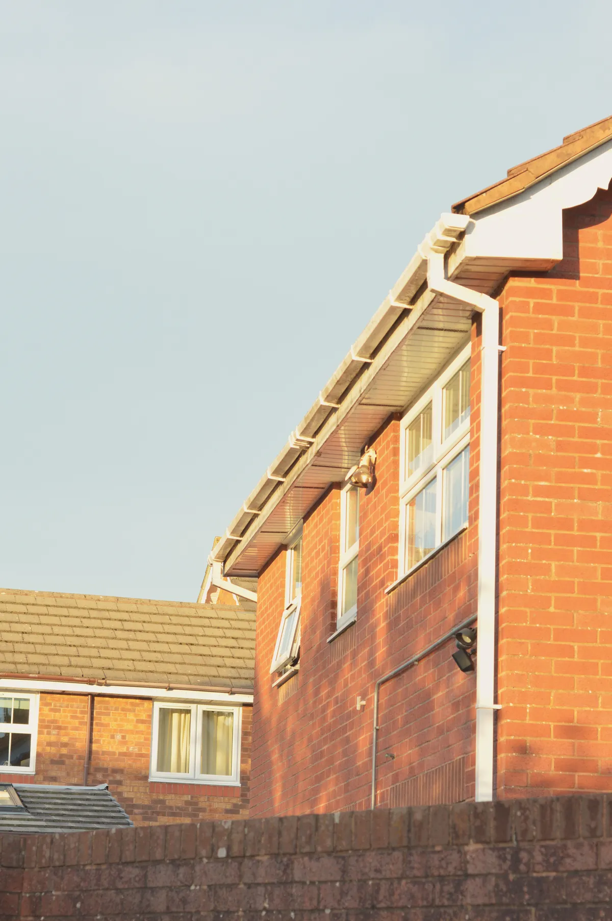 A vertical colour photograph of a red brick house. The first floor is visible. A low brick wall stops our view in the foreground of the ground floor. In the background, nearly perpendicular, another red brick house first floor is visible.