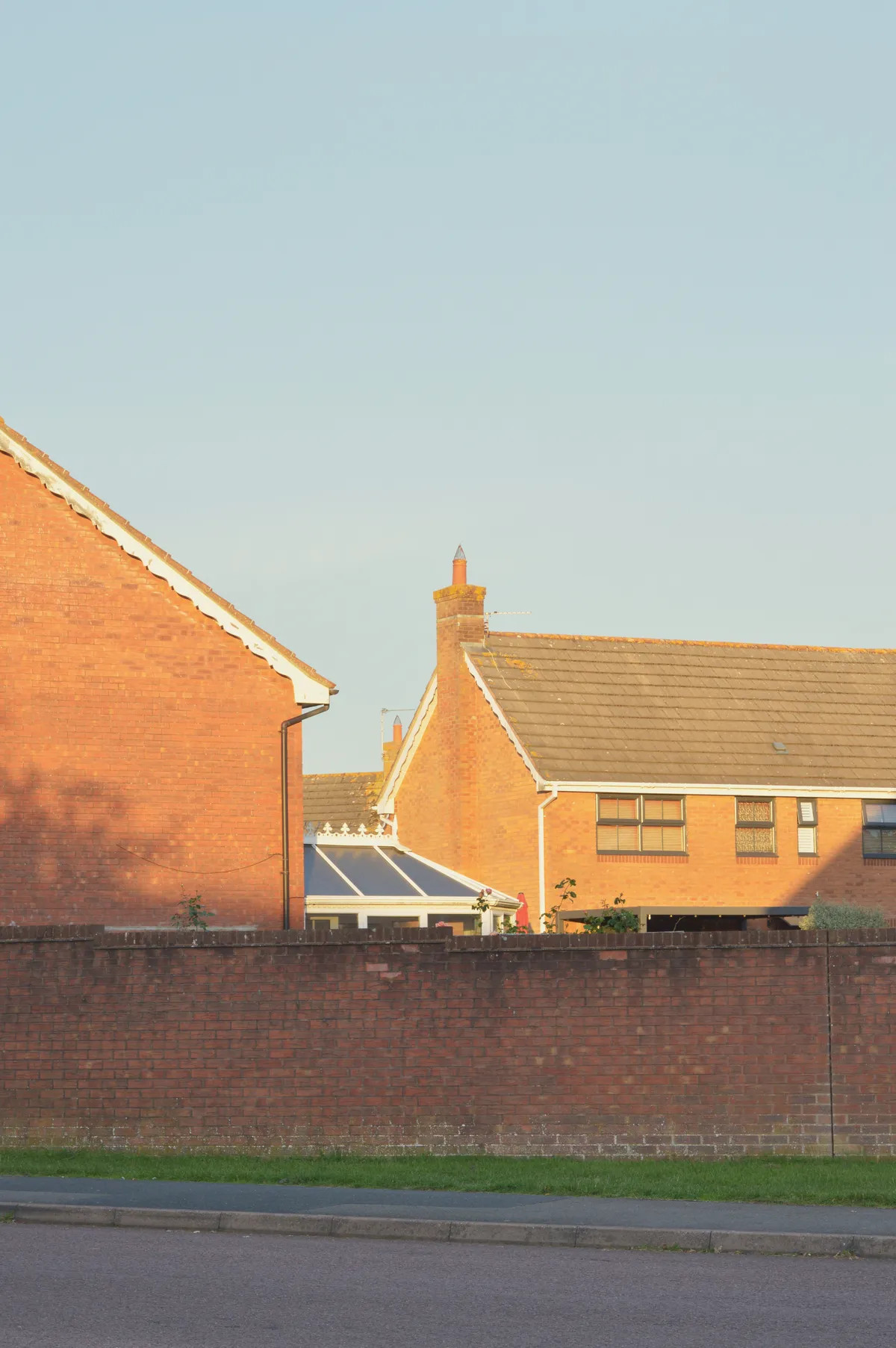 A vertical colour photograph of a street with a red brick wall in shadows. Behind the wall are red brick houses (mostly the first floors visible) bathed in golden light).