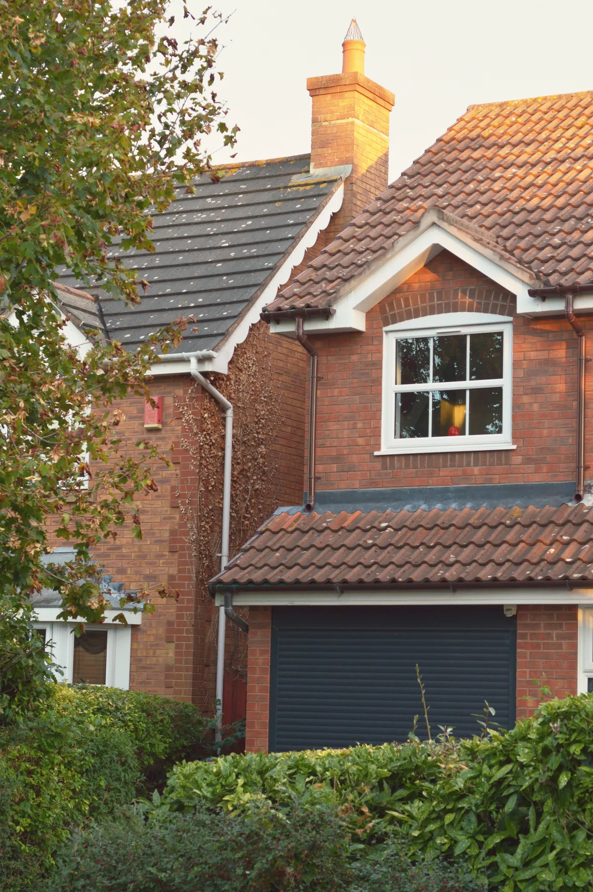 A vertical colour photograph of a red brick house surrounded by foliage. The frame is cropped so only the garage and an upstairs window are visible. The window has a shape of light reflected from the outside.