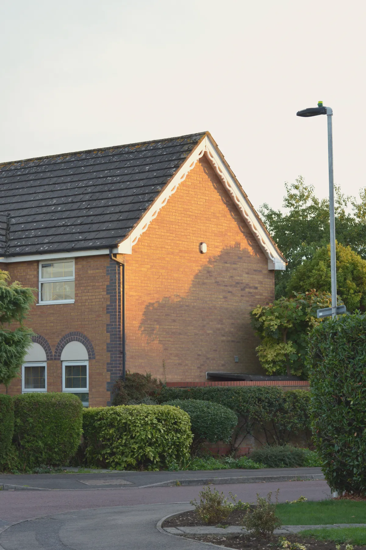 A vertical colour photograph of a turn in a street. There are green shrubs around. One red brick house is visible with a pool of golden light on a windowless wall.