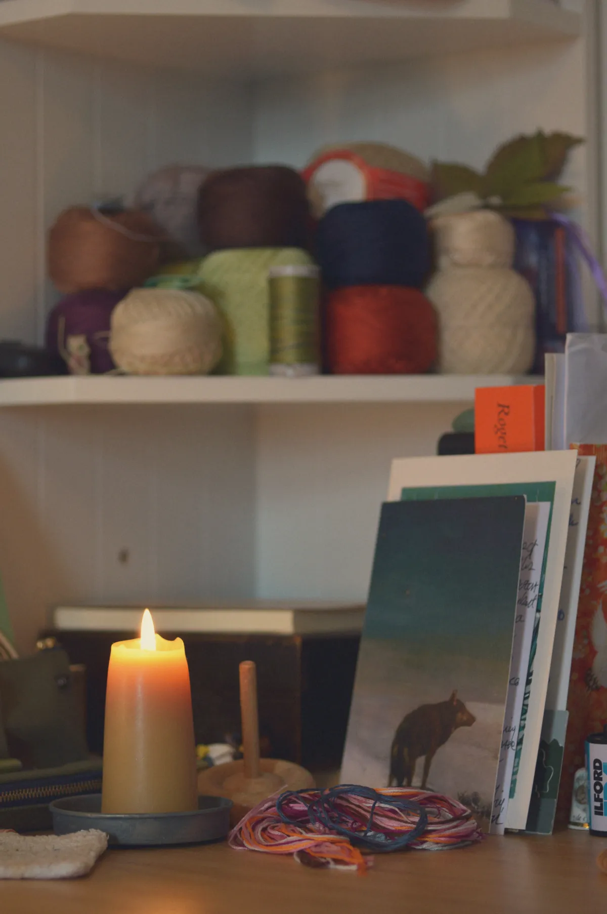A vertical colour photograph of a desk corner with a white shelving unit in the back with loads of crochet thread. In the foreground is a lit beeswax candle, a tangle of embroidery threads, and a postcard with a wolf on it.