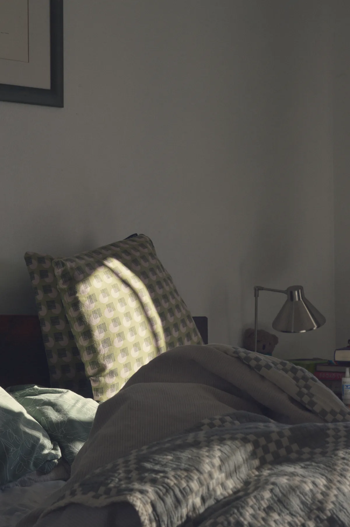 A vertical colour photograph of a corner of an unmade bed. Light bathes the cushions in the background. In the foreground a cotton cover is tangled around a duvet in a small mound in shadows.