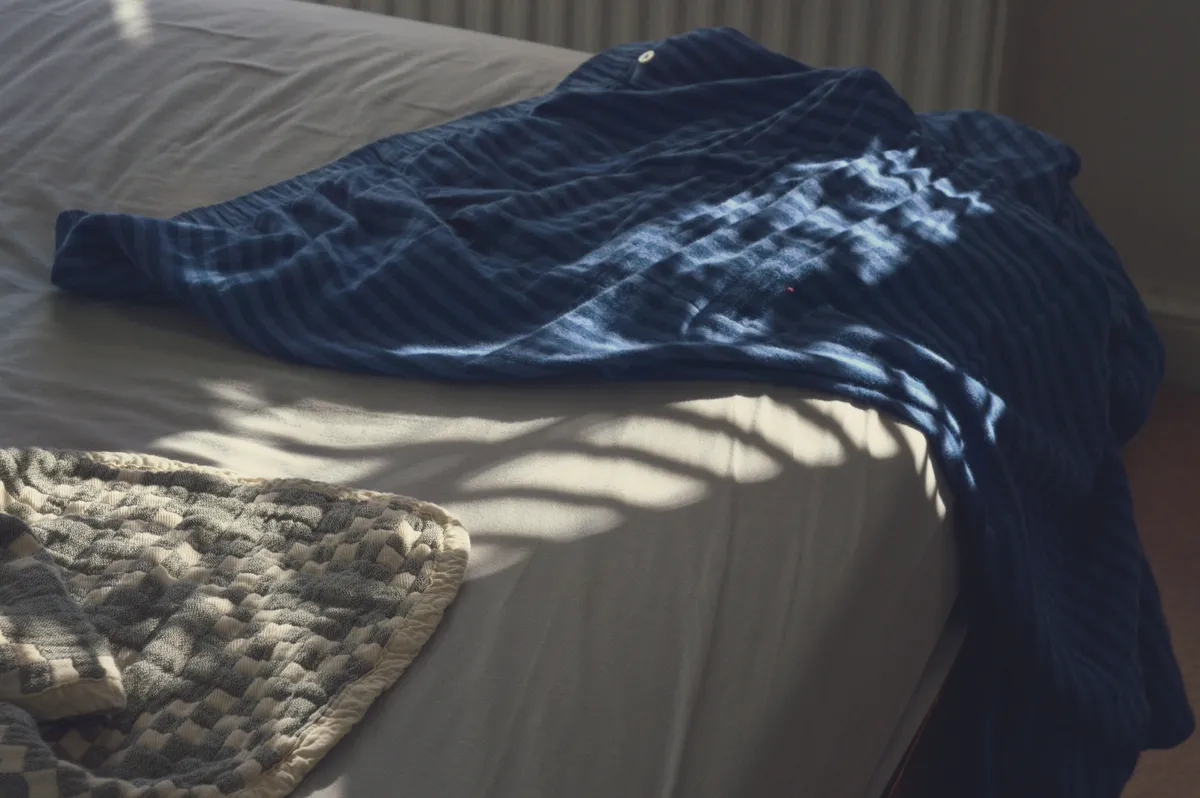 A horizontal colour photograph of a corner of a bed and the floor below. A white and grey cotton cover pokes out on the bottom right corner. A striped blue pyjama bottom hangs half off the bed on the top left. Light streaks across the scene.