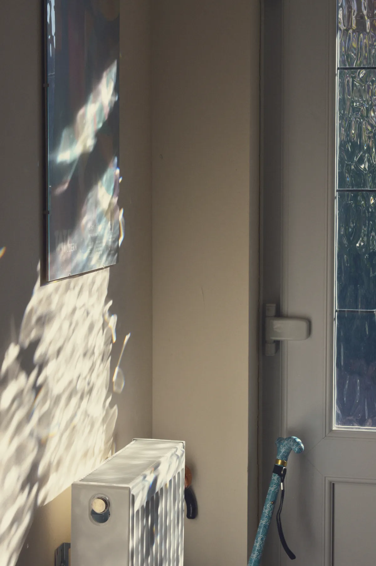 A vertical colour photograph of a corner of a house entrance. The front door is just visible in the background. The texture glass on it create ripples of light on the wall next to it. It flows down to the white radiator. On the wall is a picture frame that is mostly dark. A light blue walking stick pokes up by the door in the shadows.