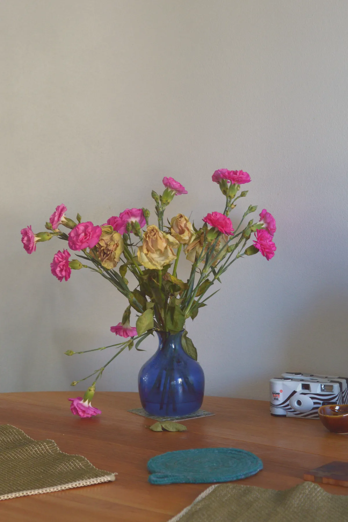 A vertical colour photograph of fading flowers - pink and white - in a blue translucent vase on a wooden table. In the foreground are green placemats and a round blue and green pot holder.