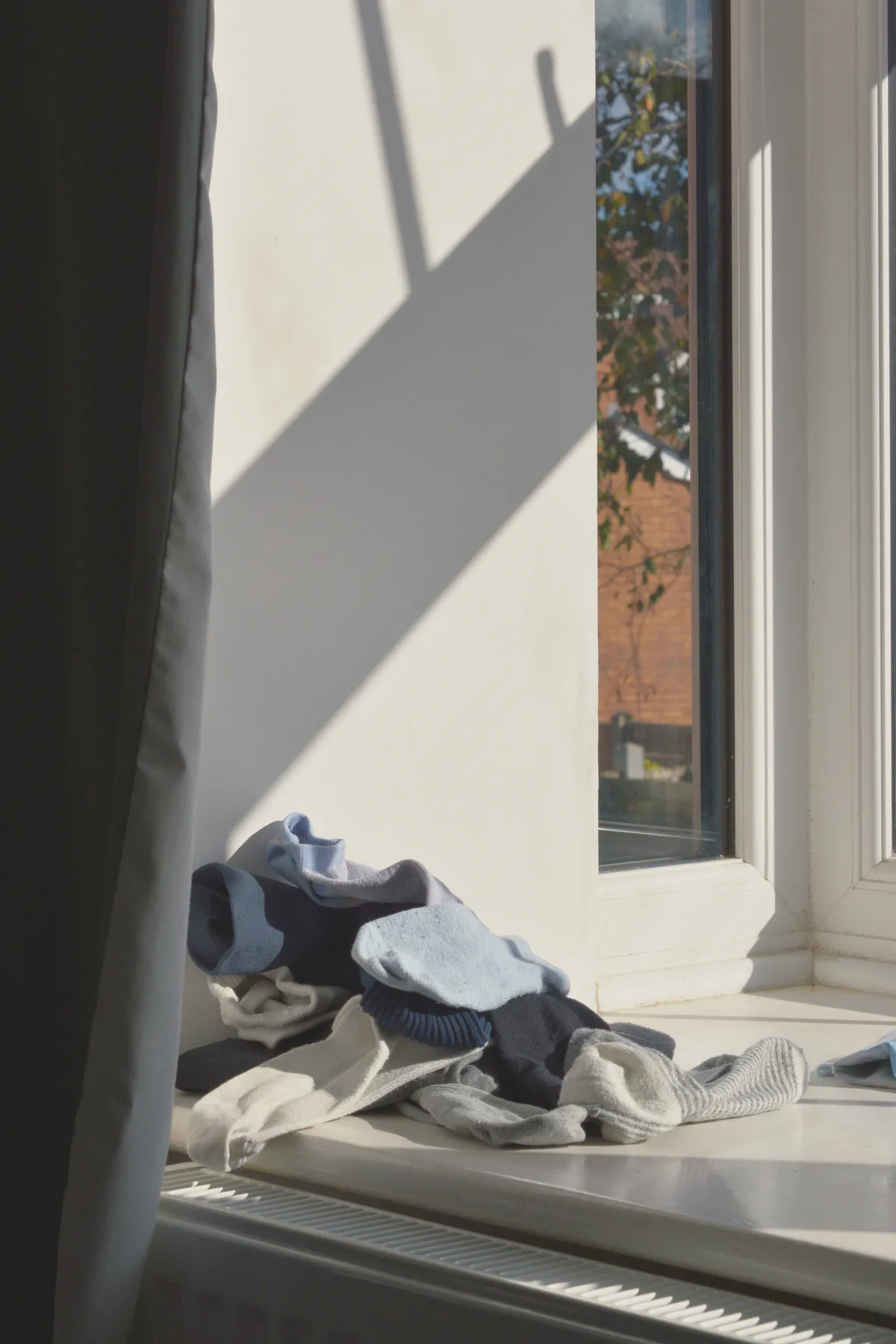 A vertical colour photograph of a pile of unmatched socks on a white windowsill. A grey curtain frames the image on the left. Light streams through from windows.