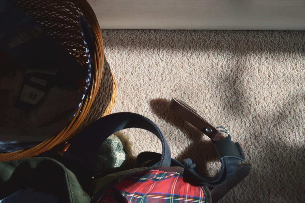 A horizontal top down colour photograph of part of a waste basket filled with paper and plastic trash. Next to it are some straps of a bag as well as part of a flap. The carpet is salmony pink. At the top of the image, a white door blocks the view.