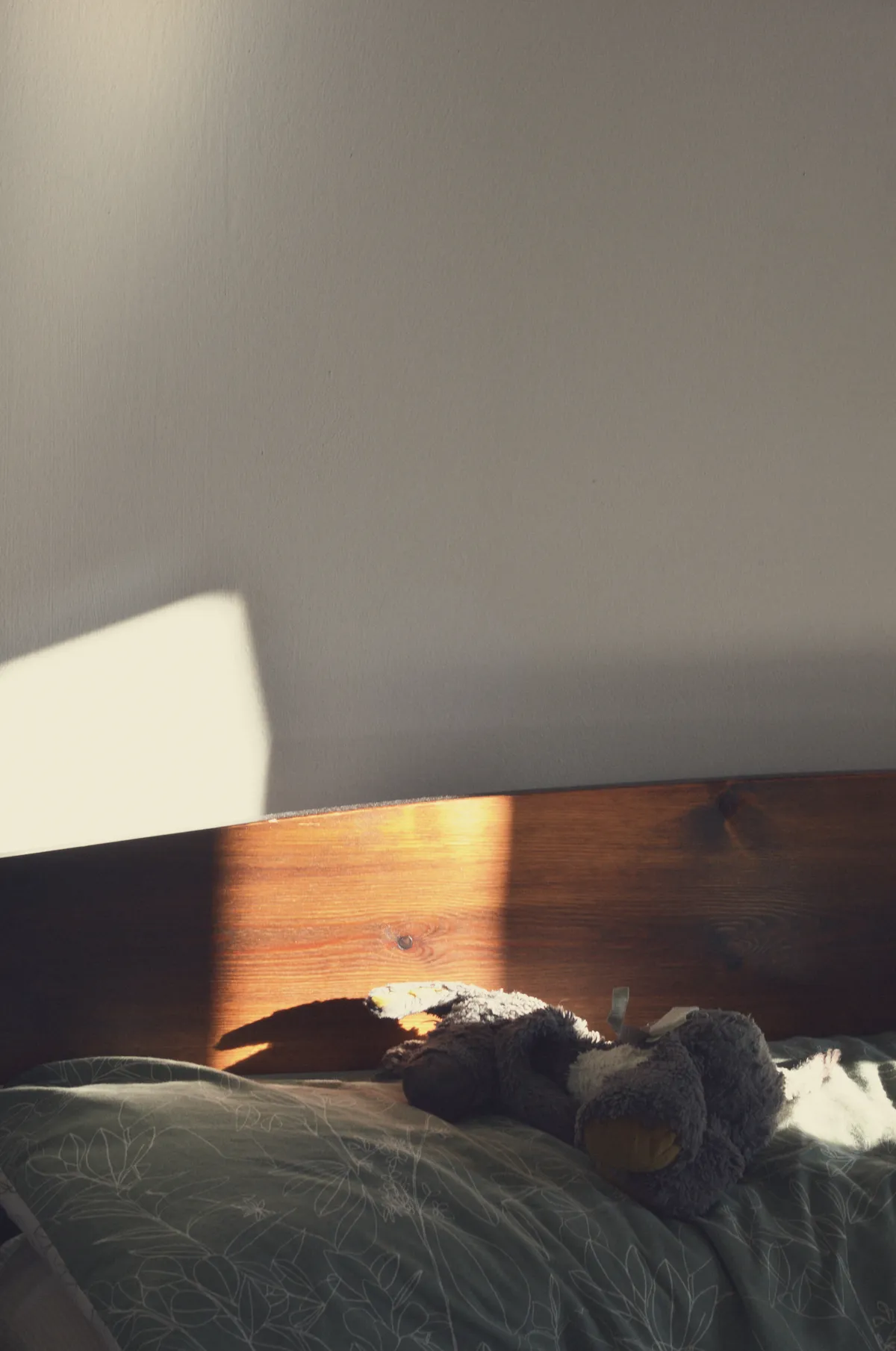 A vertical colour photograph of a corner of a bed headboard. The wood is dark brown. The wall behind is white. On the bed is a green and white floral pillow with a grey donkey plushie lying on their side. A rectangular streak of light illuminates part of the scene, including some of the donkey.
