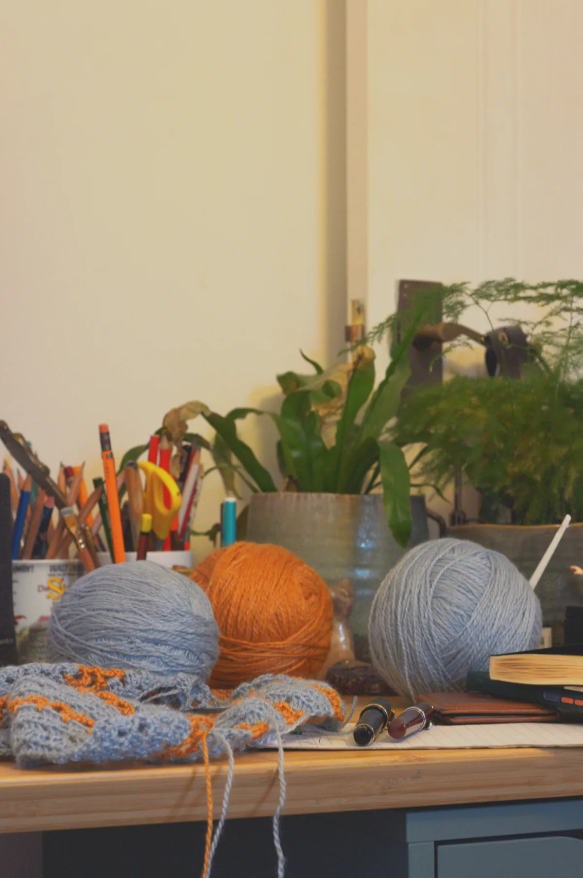 A vertical colour photograph of three balls of wool (two light blue and one orange) on a desk with a crochet work in progress in front of them. Next to the work are two fountain pens. Behind are some plants, mugs with desk paraphernalia, and other bits and bobs.