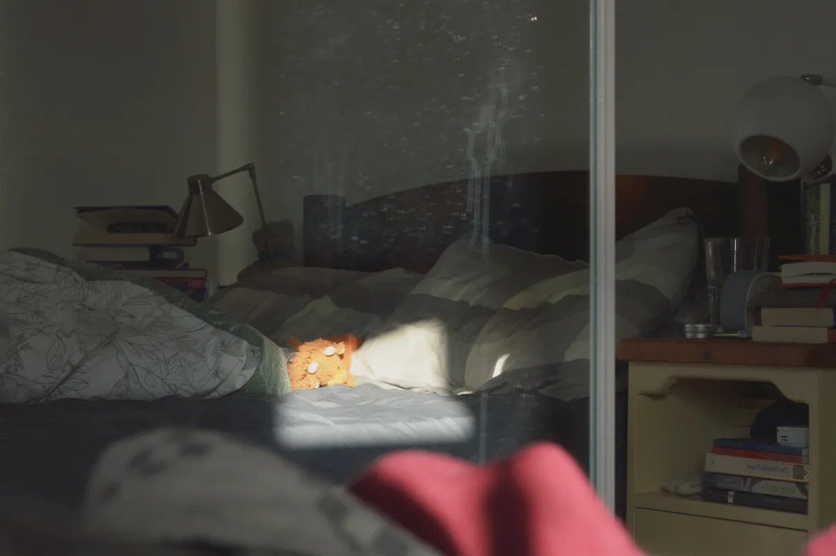 A horizontal colour photograph of a highland cow plushie reflected in a mirror in the midst of an unmade bed. The mirror is dirty.