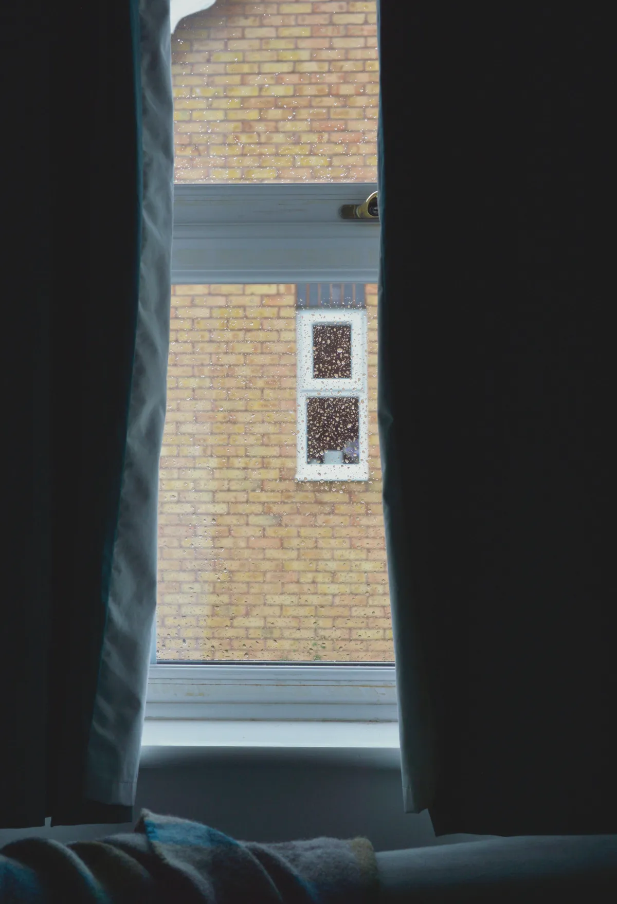 A vertical colour photograph of a window splattered with rain drops and framed by dark teal curtains. The top of a grey sofa with a wool blanket on it is just visible. The view through the window is of a yellow brick house.