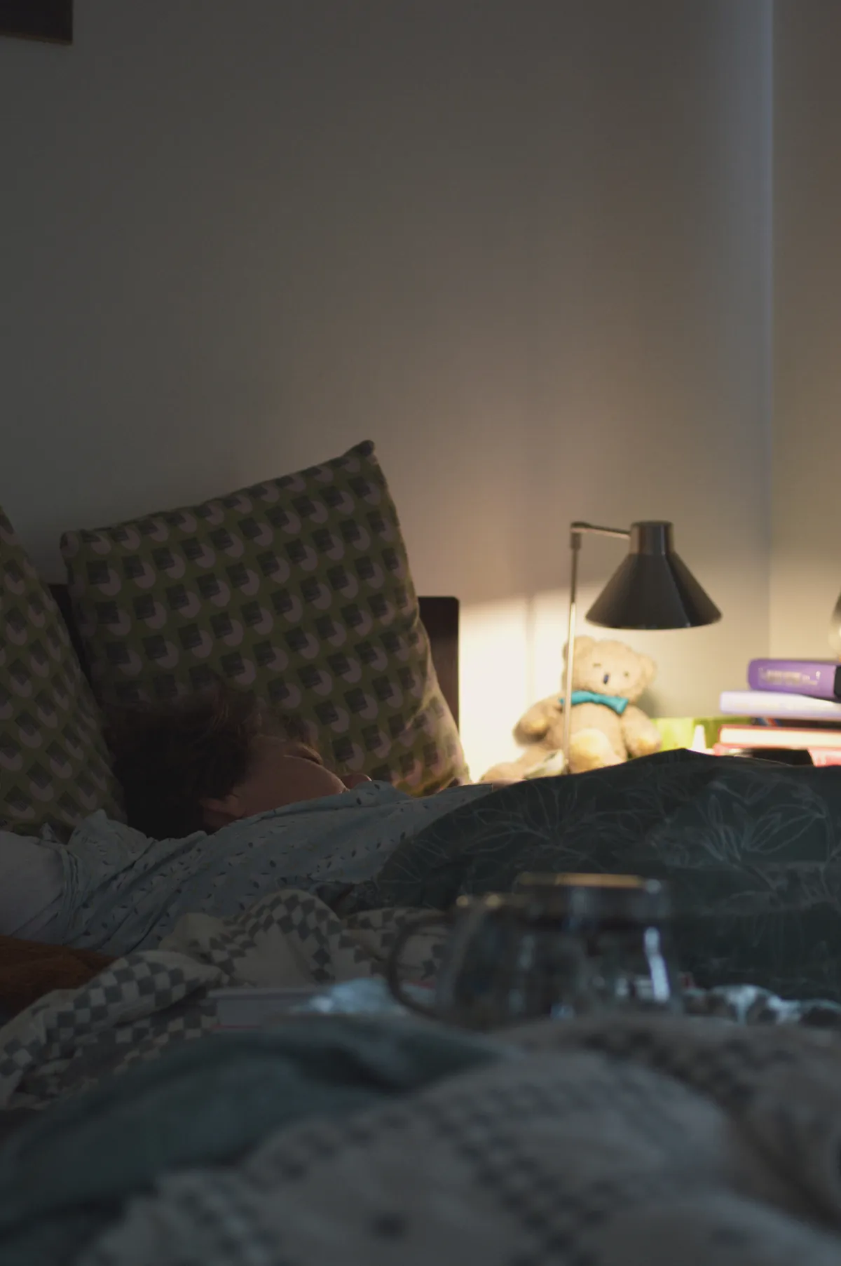 A vertical colour photograph of a bed and bedside table. In the bed a person is asleep, their face turned away from the camera. On the bedside table, a light shines on a teddy bear. The rest of the scene is darker.