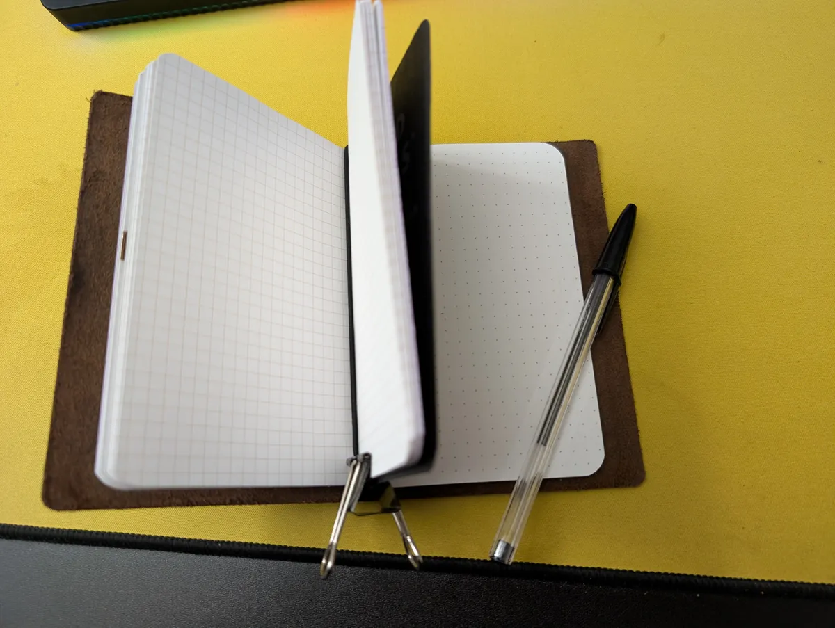 Two Field Notes notebooks, one graphed and the other dotted, held together in a leather cover and help open with a binder clip. A black Bic Crystal ballpoint pen sits inside the second notebook