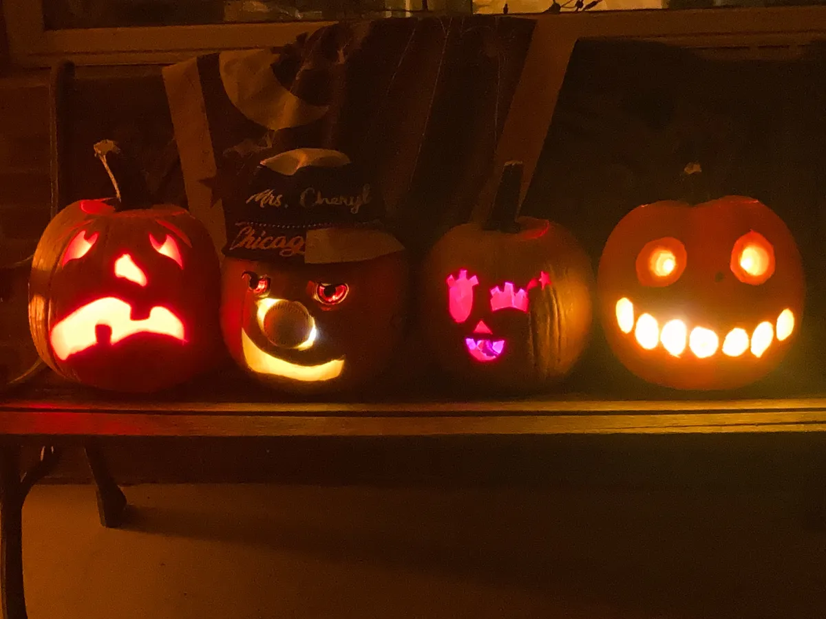Four jack-o-lanterns sitting on a front porch bench at night, with their lights on