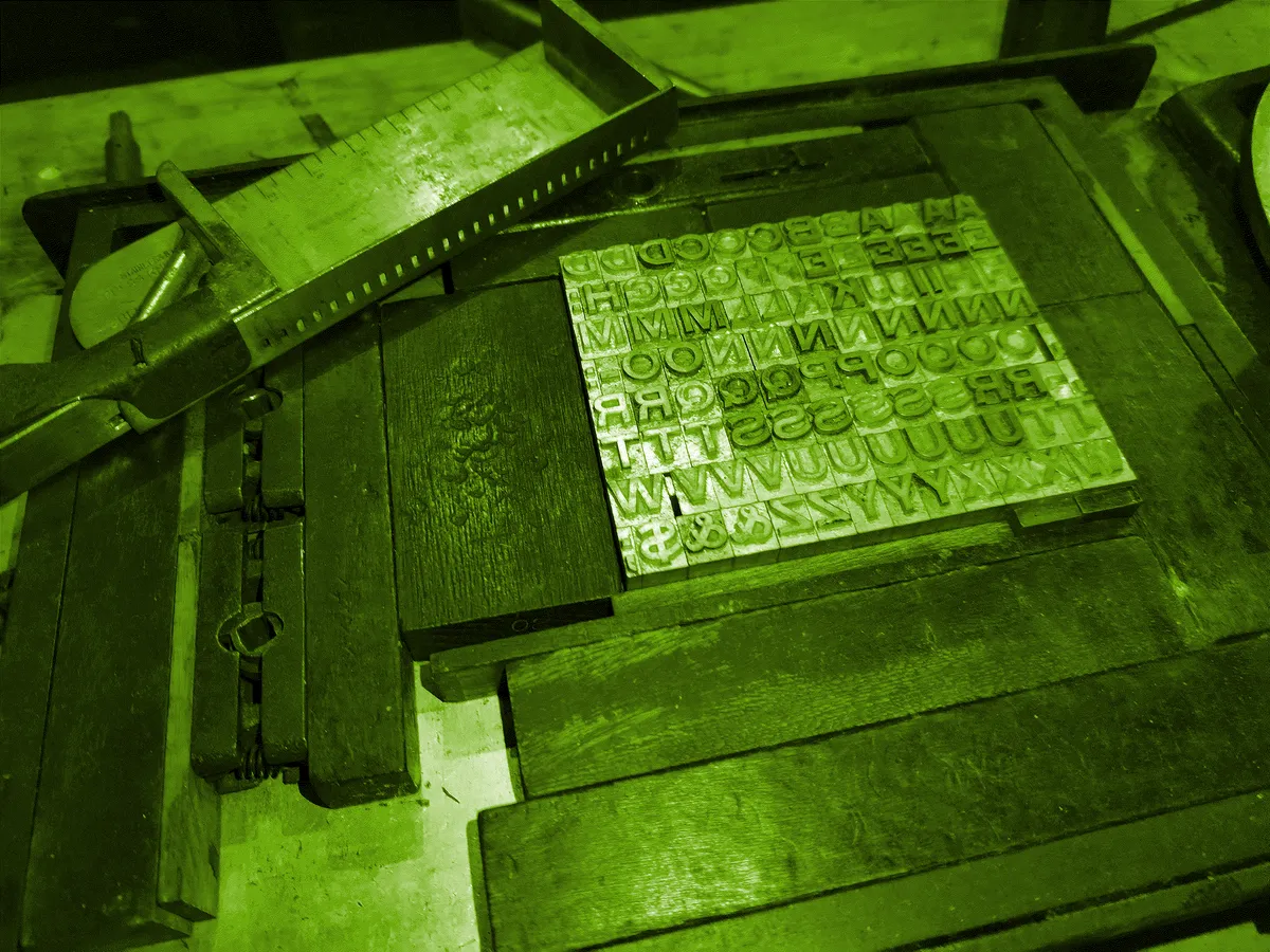 green-tinted close-up of a letterpress work surface: composing stick, ruler, and a grid of metal sorts.