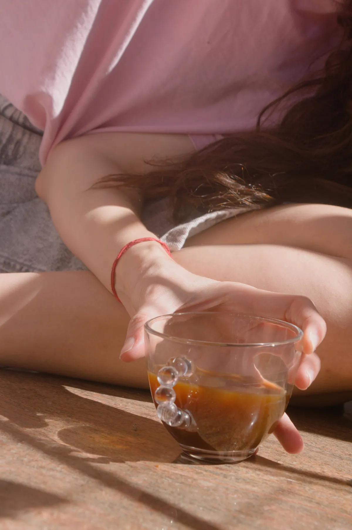 My girlfriend having a cup of coffee on my tiny balcony. She's wearing a pink t-shirt and gray denim shorts. The coffee cup is made of clear glass. The photo is shot close up to her hand holding the coffee cup on the ground. Other than her hand, legs, clothes, and a little bit of her hair, everything else is kept out of the shot.