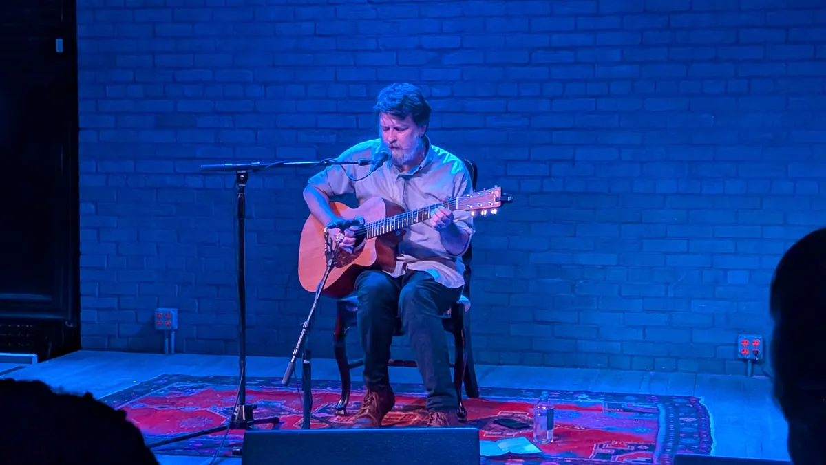 A man sitting with a guitar, playing before a crowd in a pastoral venue.