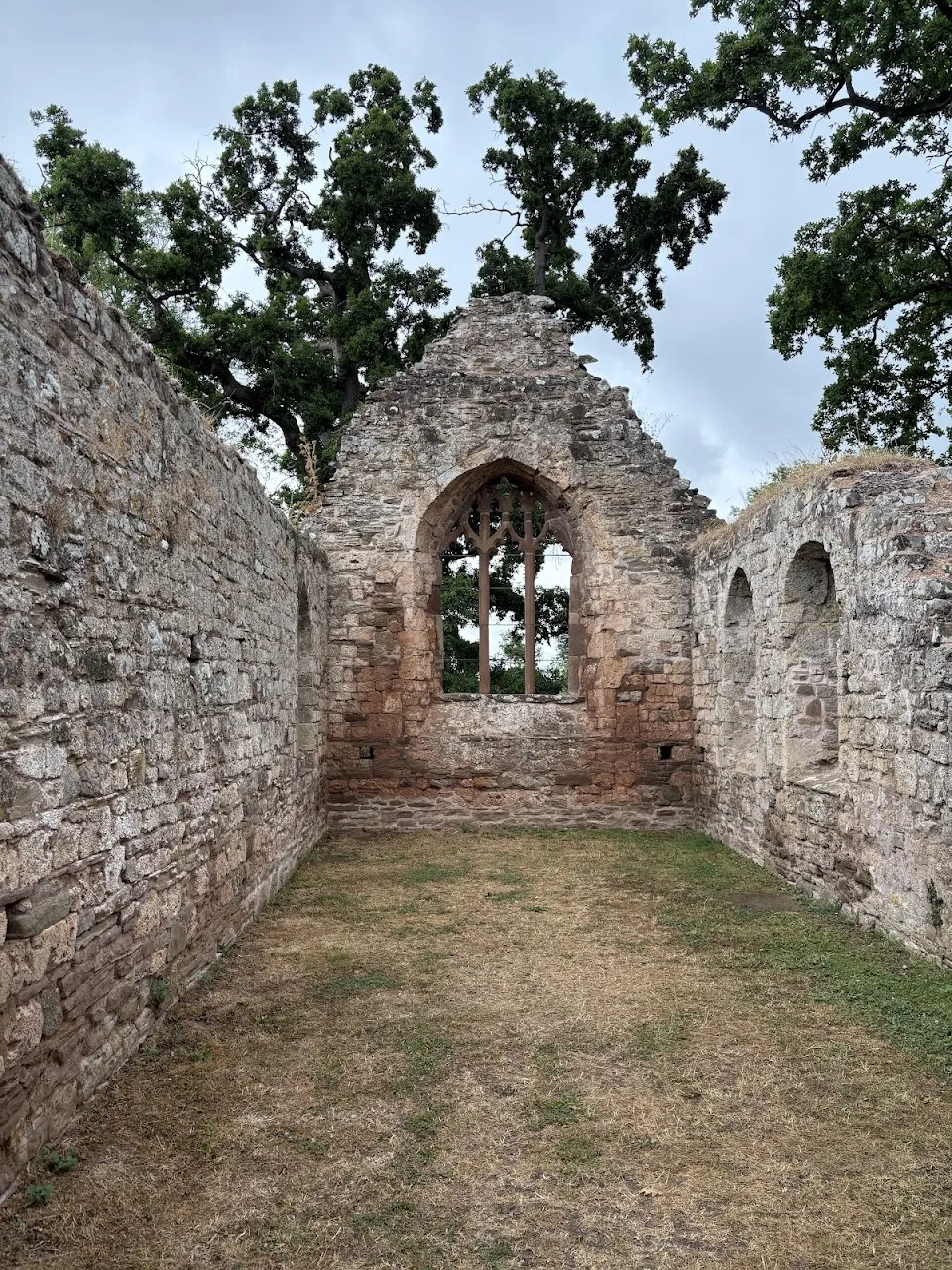 Interior of roofless stone ruin with pointed arch window framing trees and cloudy sky; dry grass on floor