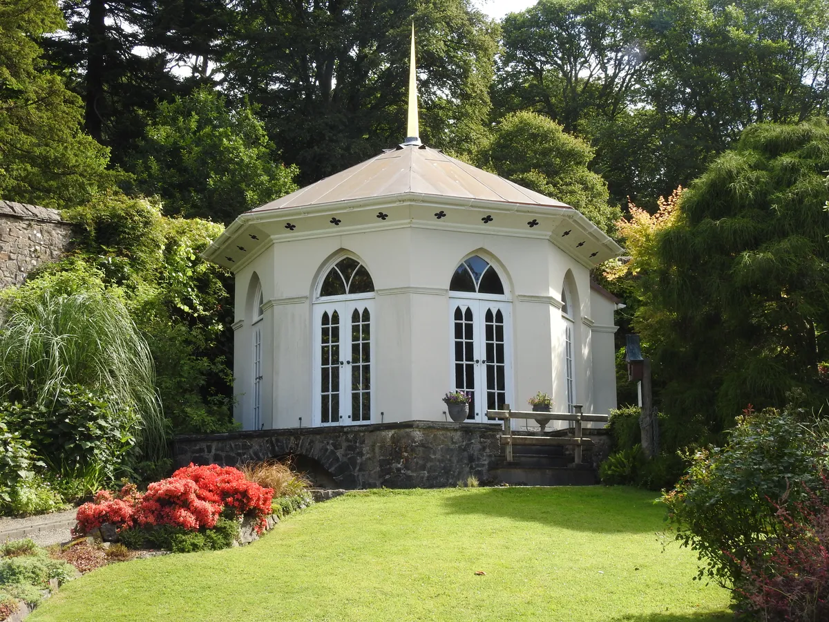 The Victorian Gothic gazebo at the heart of Colby's walled garden, an octagonal building with a copper cone-shaped roof crowned by a golden gilded pinnacle. Pointed Gothic windows and a pair of glazed doors look out over a neatly mown lawn, with a vivid clump of red azalea flowering beside the stone terrace. Inside are trompe l'oeil murals by the artist Lincoln Taber.
