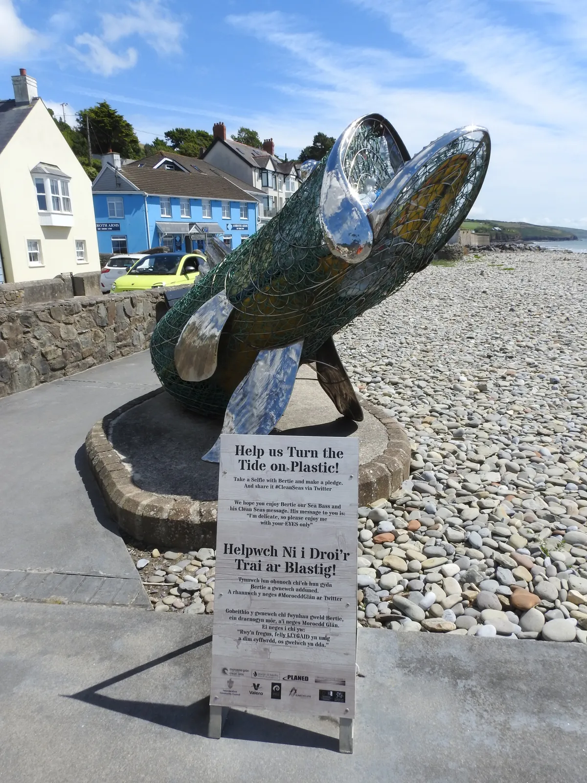 A striking sculpture of a sea bass, woven from wire mesh and polished metal, stands on the seafront at Amroth beside a 'Turn the Tide on Plastic' pledge board in English and Welsh, raising awareness of ocean plastic. Behind it the pale blue Amroth Arms and neighbouring cottages line the road. The pebble beach and headland stretch away to the right.