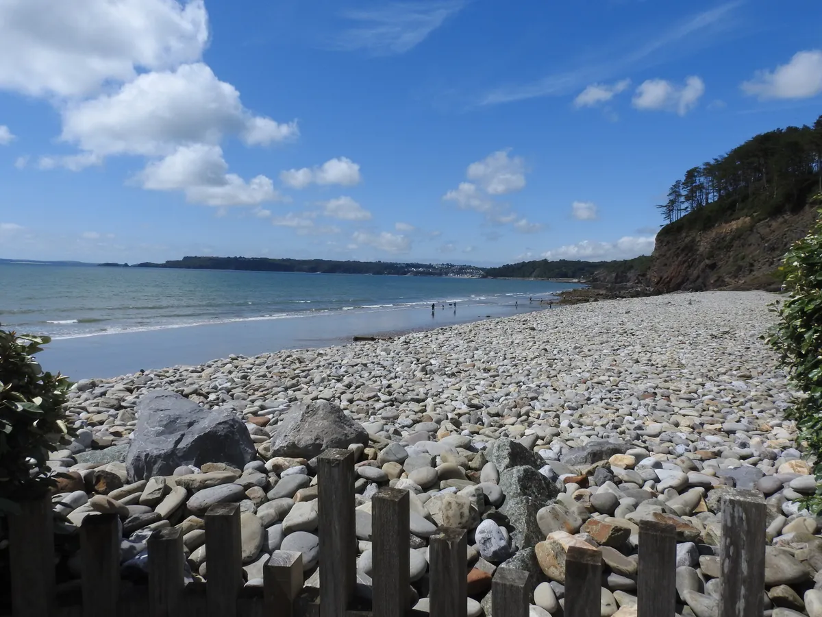 Looking out from Amroth along the pebble beach towards Saundersfoot, the coastline curving away beneath a wooded headland. A bright blue sky scattered with fluffy cloud sits above a calm sea, with a few figures paddling at the water's edge. A weathered wooden fence and large boulders frame the foreground.