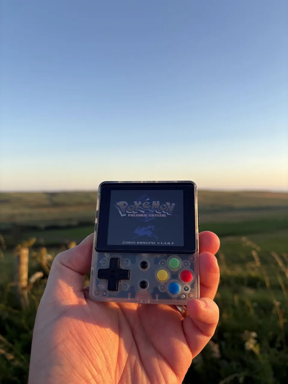 Hand holding transparent gaming device showing Pokémon Polished Crystal title screen; grassy field and sunset sky in background