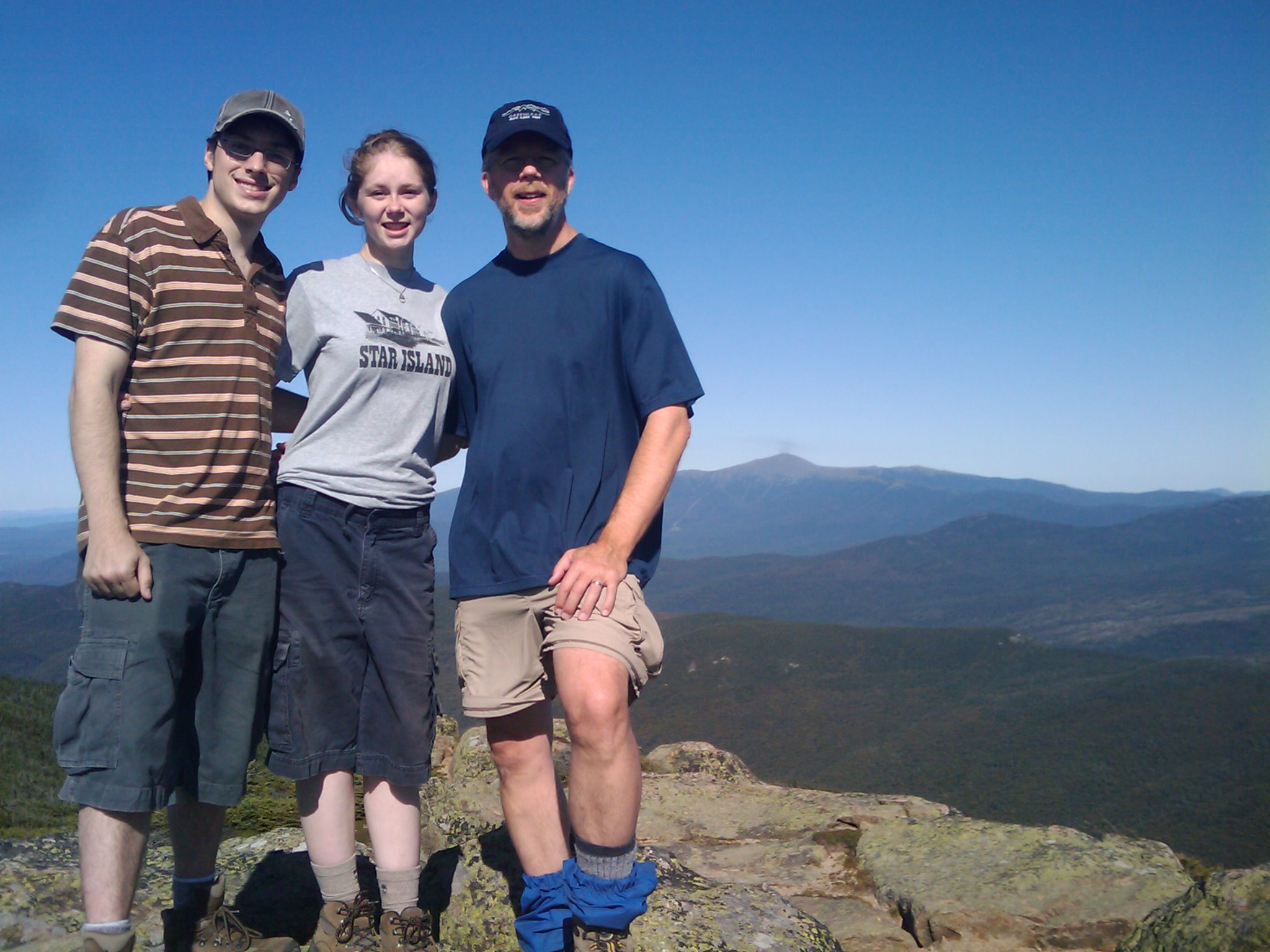 Three hikers at the peak of a mountain with several mountains in the background, and clear blue skys. The hiker on the right is a middle aged man, with a short beard, and is dressed in a blue shirt with a blue baseball cap from one of the nearby huts. The hiker in the middle is a young woman wearing a gray star island t-shirt and long blue shorts. The hiker on the left is a young man wearing a gray 2K baseball cap with brown and orange striped shirt and long blue shorts, he's clean shaven and wearing glasses. They're smiling and looking very relaxed.