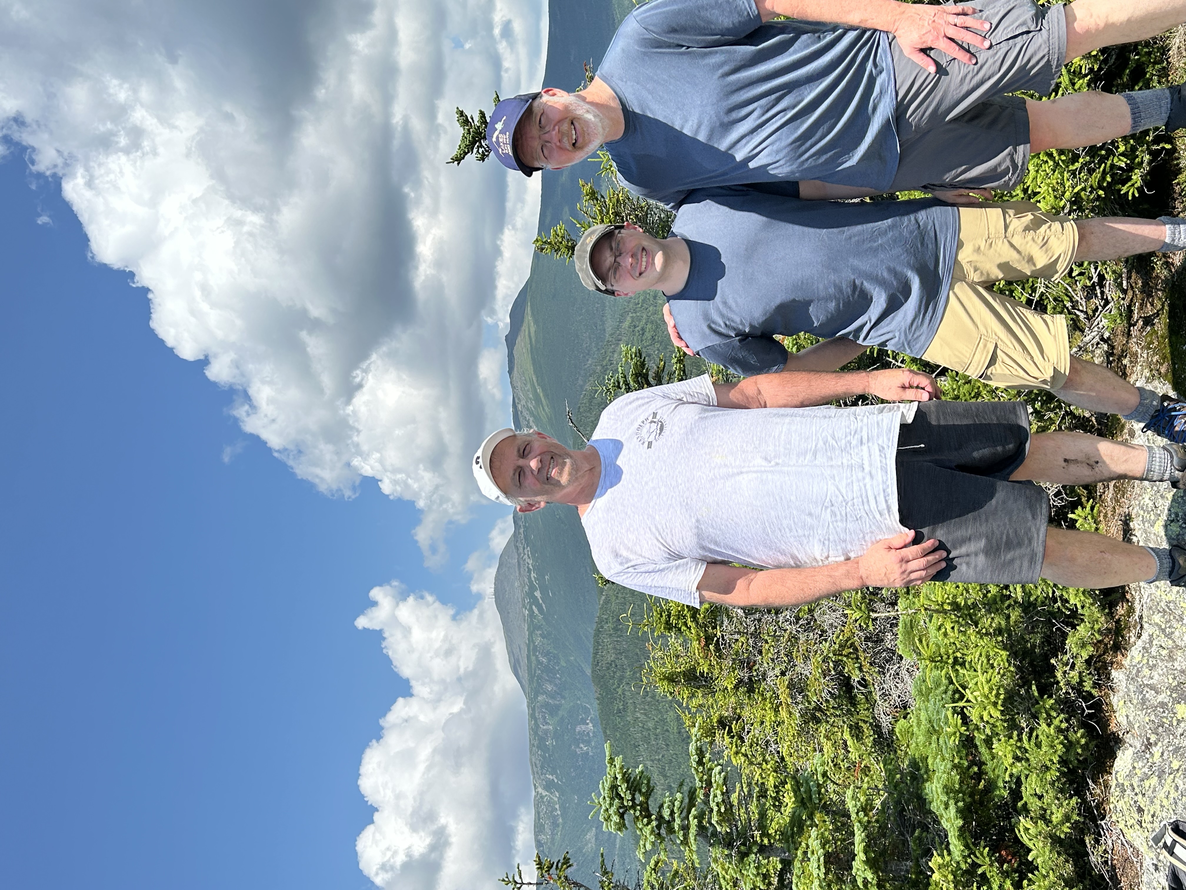 Three hikers at the peak of a mountain with several mountains in the background, and large white clouds. The hiker on the right is an older man, with a short grey beard, and is dressed in a blue shirt with a blue baseball cap from one of the nearby huts. The hiker in the middle is a middle aged man wearing a gray 2K baseball cap with a blue shirt and tan shorts blue shorts, he's clean shaven and wearing glasses. The man on the left is an older man, clean shaven, with a white baseball cap and white shirt. They're smiling and looking very relaxed.