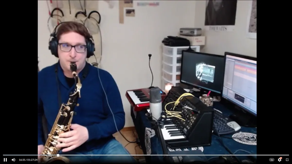 A person playing saxophone. A desk is to his left, housing synthesizers and computer monitors