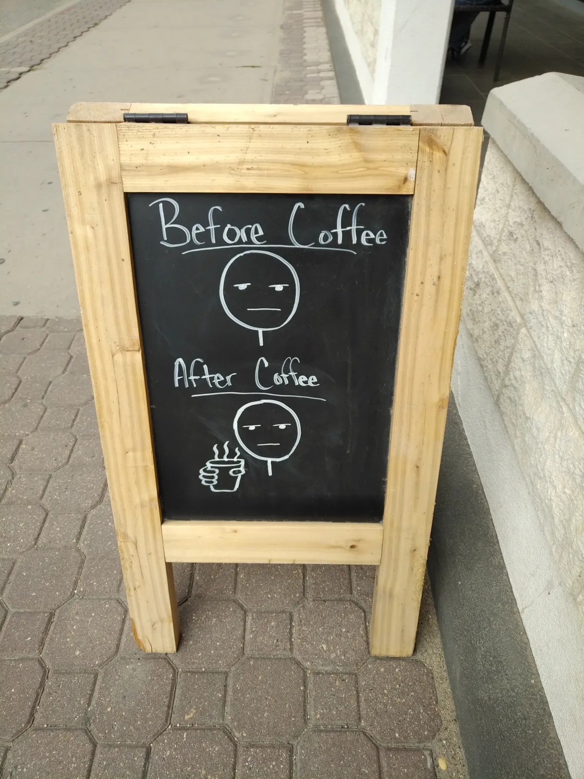 A sidewalk sandwich board showing an expressionless face labelled "Before Coffee" and another expressionless face with a coffee cup beside it labelled "After Coffee"