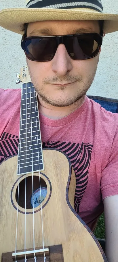 A person leaning back, wearing a hat and sunglasses, a ukulele resting across his torso and over his shoulder