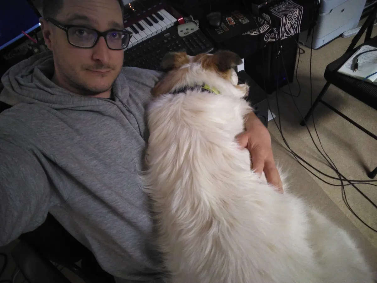A person with a large dog in his lap. A desk with a piano keyboard and computer keyboard can be seen in the background.