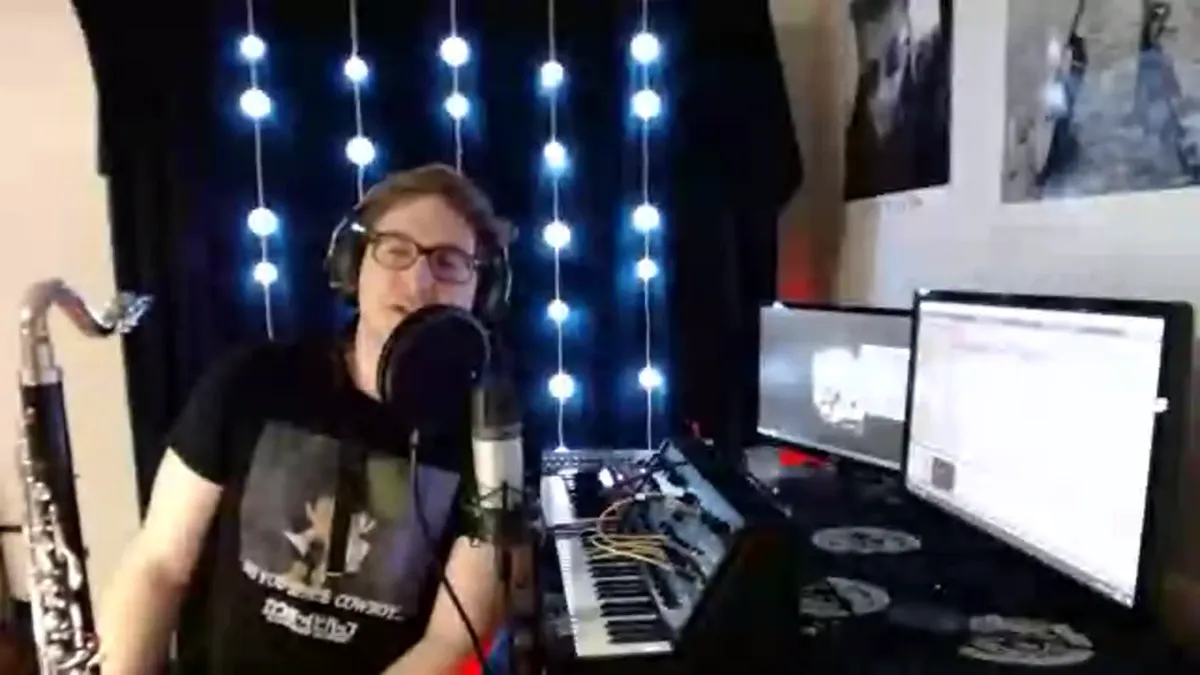 A person holding a bass clarinet and singing into a microphone. A synthesizer and computer monitors sit on a desk beside him.