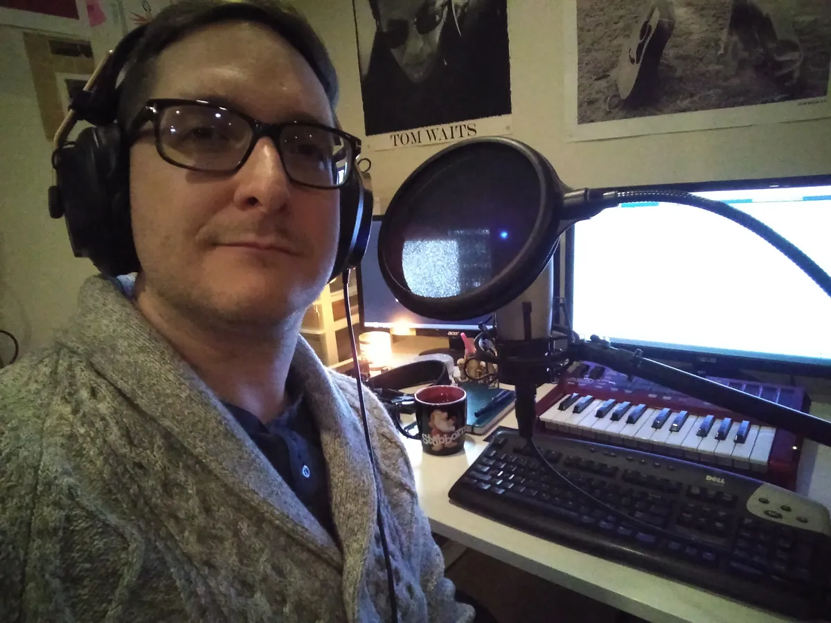 A person wearing headphones in front of a mic with a pop screen. A desk with monitors, a computer keyboard and a midi keyboard are in the background