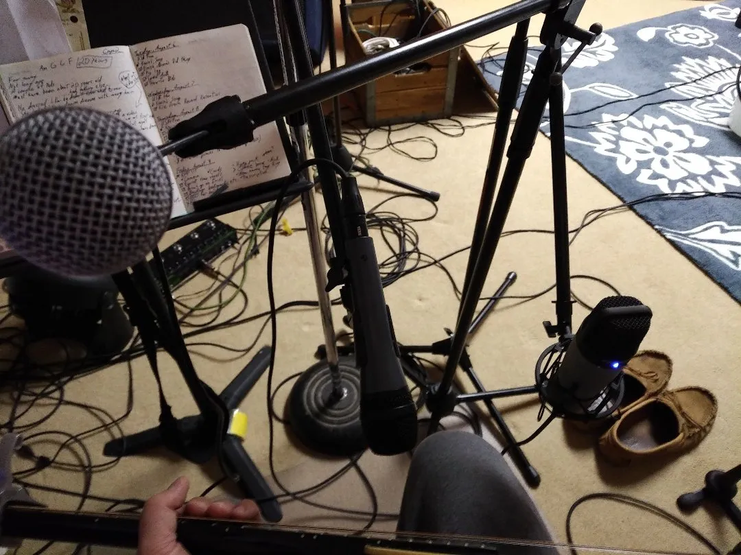 The photo is taken from the perspective of someone sitting down, holding a guitar. There are three mics facing the photographer and a mess of cables on the floor. A notebook on a music stand is in the background