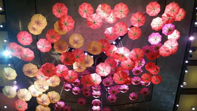 Multicoloured umbrellas, hanging, upside down, from a high ceiling, backlight by the ceiling lights, resembling flowers.